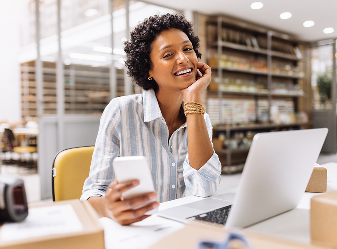 Mulher sorridente usando smartphone e laptop em ambiente de trabalho moderno, transmitindo alegria e produtividade.
