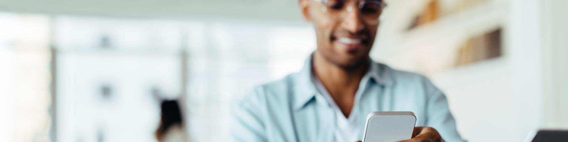 Homem usando smartphone ao lado de um notebook, em ambiente de trabalho moderno, simbolizando tecnologia financeira, open finance e gestão digital de finanças