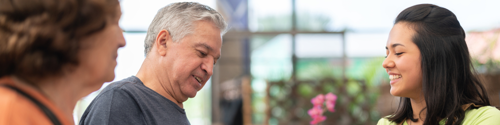 Homem idoso sorridente conversando com duas mulheres em uma loja, ambiente iluminado e acolhedor.